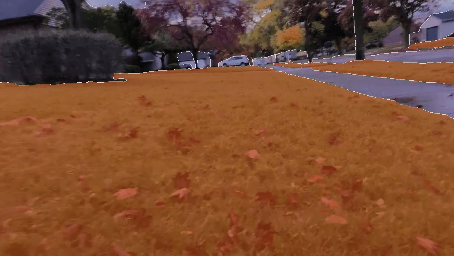A suburban street in autumn with orange-tinted grass and trees, fallen leaves scattered on the ground, resembles scenes captured in the KITTI dataset used for Semantic Segmentation.