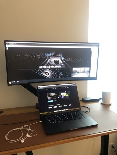 Workspace featuring a laptop and a monitor displaying 3D mapping data, with earphones and a coffee cup on the desk.