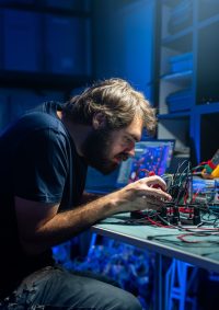 A man working intently on electronics at a desk, surrounded by wires and equipment, in a dimly lit room with blue lighting.