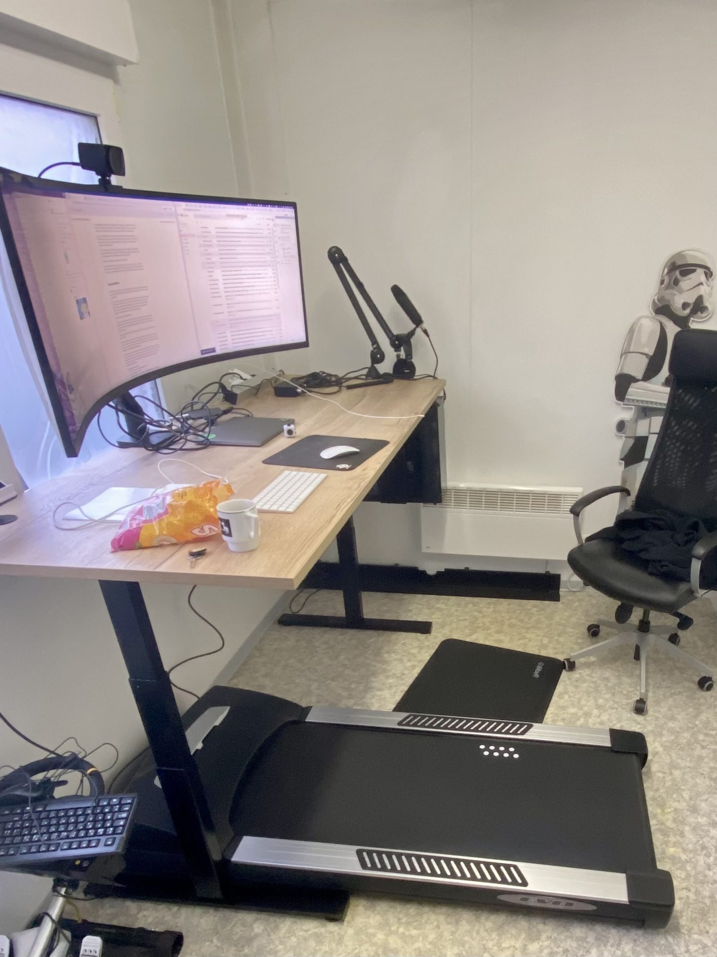 Justin engineering workspace Home office with a standing desk, monitor, treadmill, chair, and a cardboard cutout. Snacks and a mug are on the desk.
