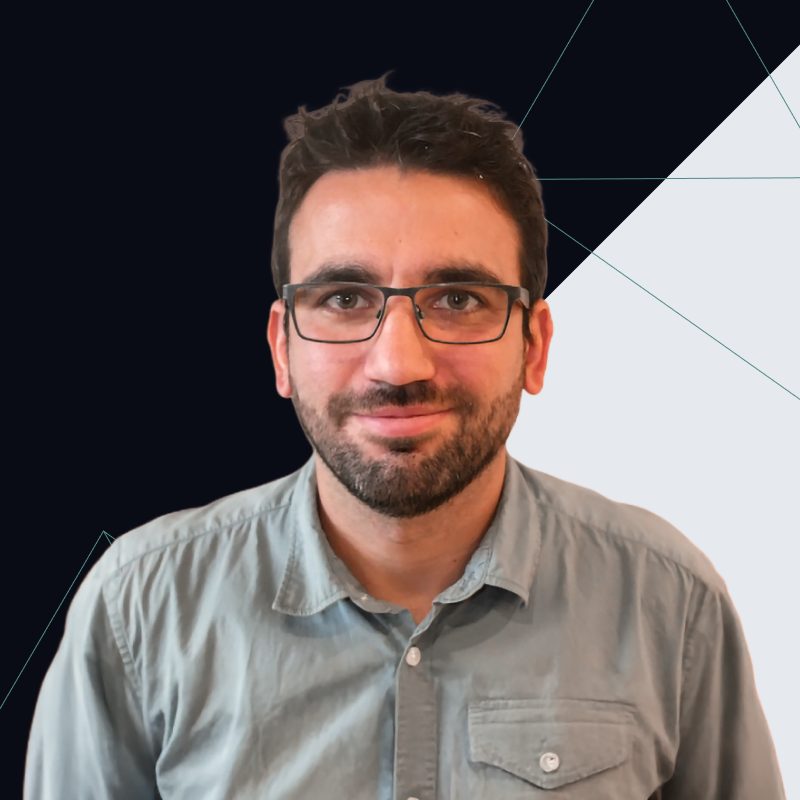 Man with glasses and a beard wearing a light gray shirt, standing against a dark geometric background.