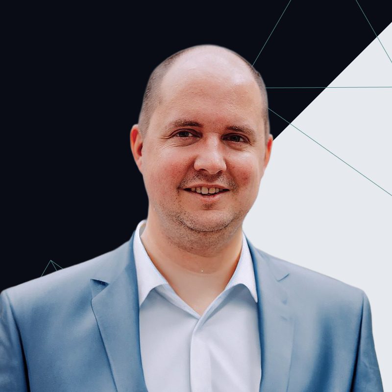 A smiling man in a light blue suit and white shirt stands against a geometric, dark and light background.