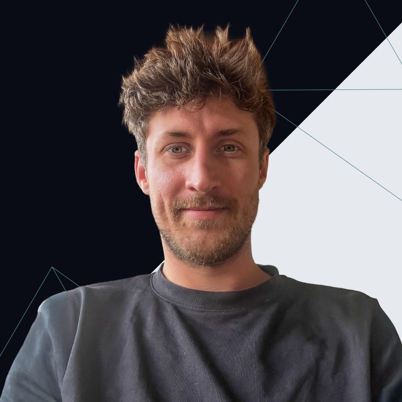 A man with wavy brown hair and a beard smiles at the camera against a modern geometric background.
