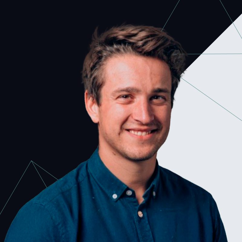 A man with short brown hair smiles, wearing a blue shirt against a black and white geometric background.
